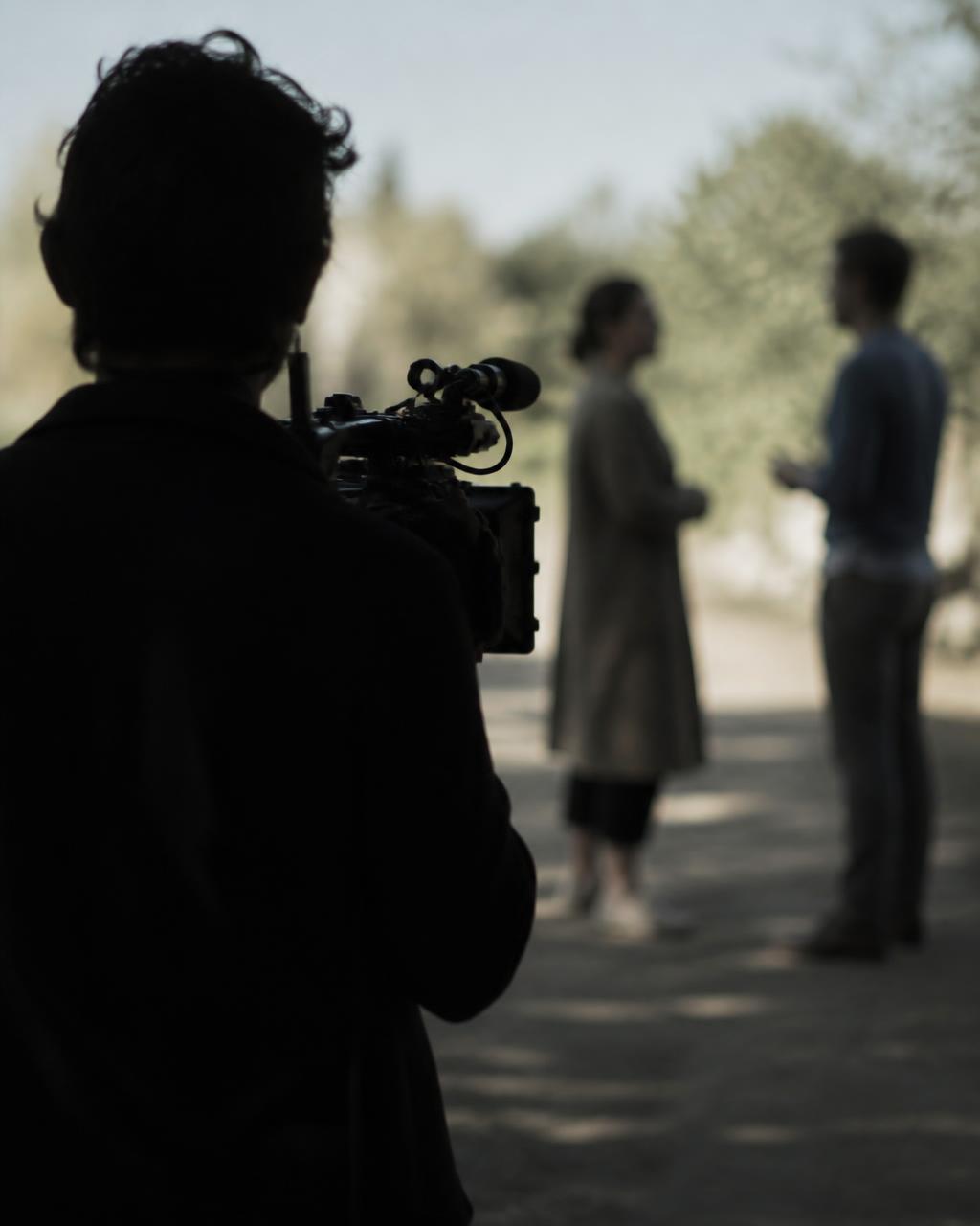 A camera operator seen from behind, observing two people talking in daylight